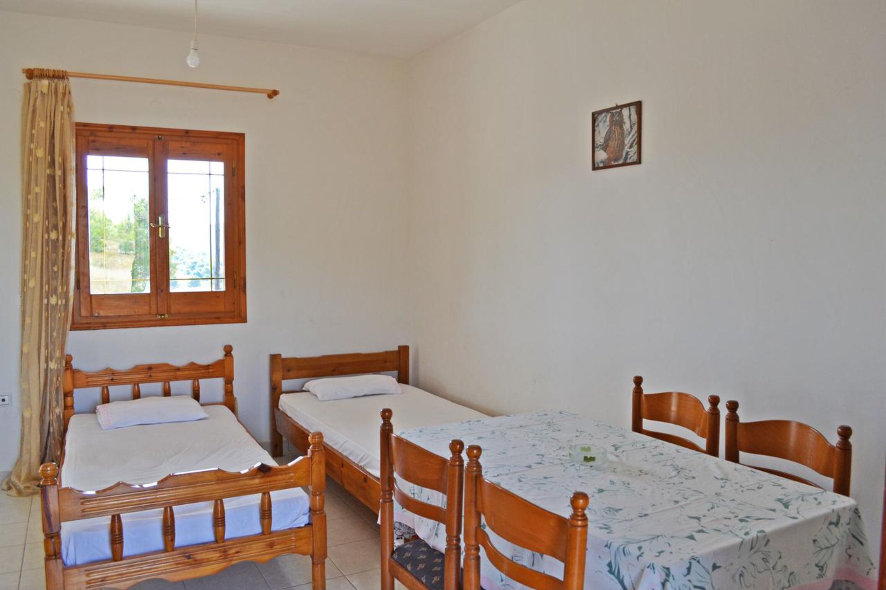 A room with two wooden beds, a dining table with chairs, a window with curtains, and a small picture on the wall.