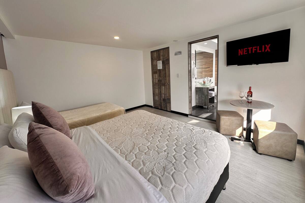 Ein Hotelzimmer mit einem Bett, einem Fernseher, einem kleinen Tisch mit einer Flasche und Gläsern sowie einem durch eine offene Tür einsehbaren Badezimmer.