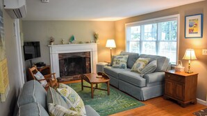 A living room with a fireplace, a sofa, a television, and a window. - Lovely three bedroom home with access to Swan Pond (Dennis Port)