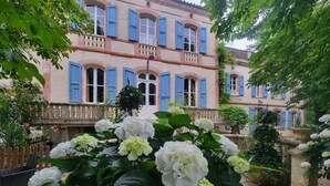 Un edificio con múltiples ventanas y balcones, rodeado de abundante vegetación y flores blancas.