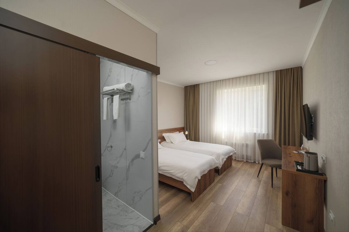 A hotel room with two beds, a wooden desk, a chair, a TV, and a shower area.