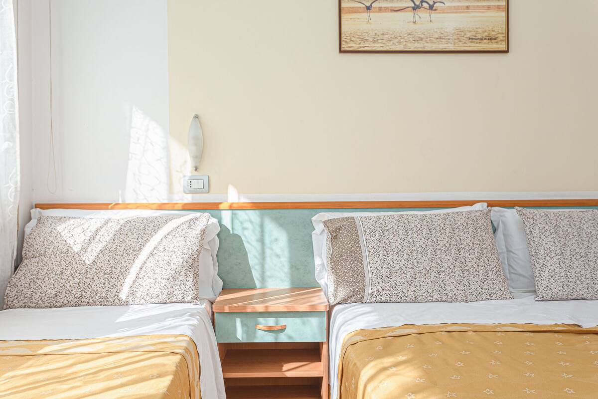 A bedroom with two beds, a nightstand, a wall-mounted light, and a painting.
