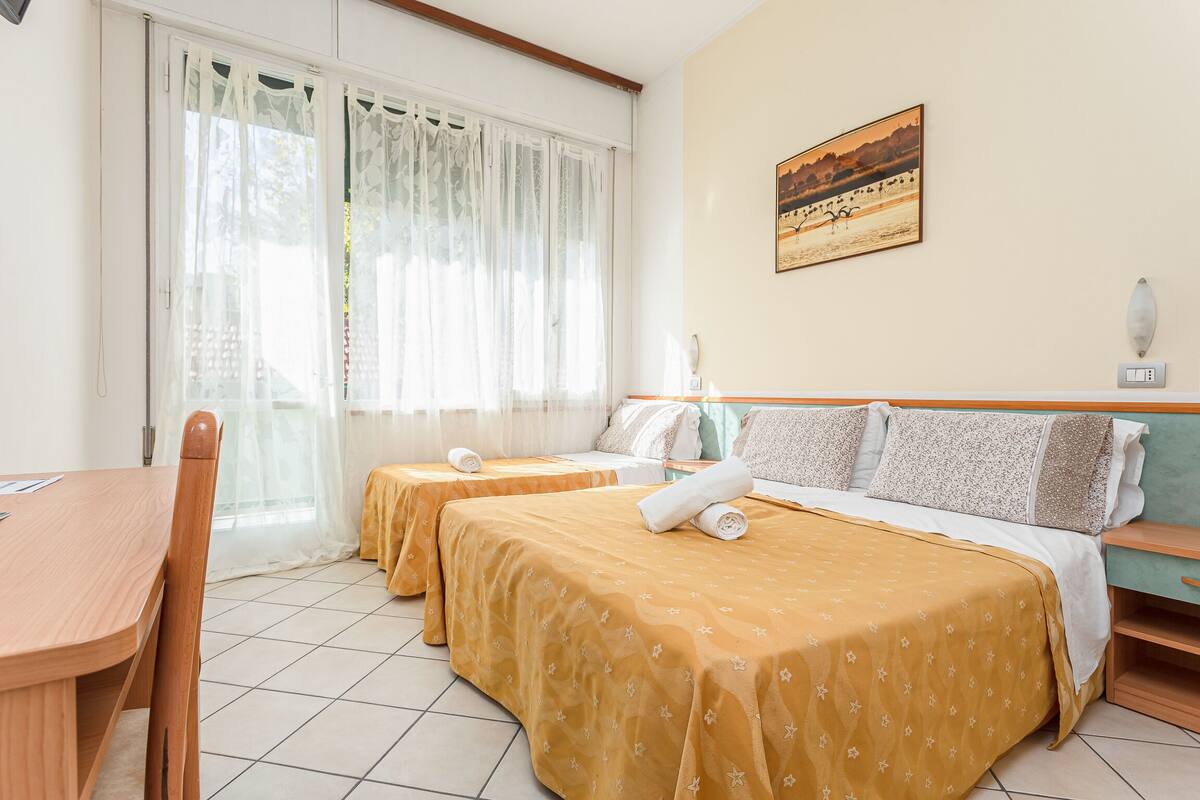 A hotel room with two beds, a wooden table, and a window with sheer curtains.