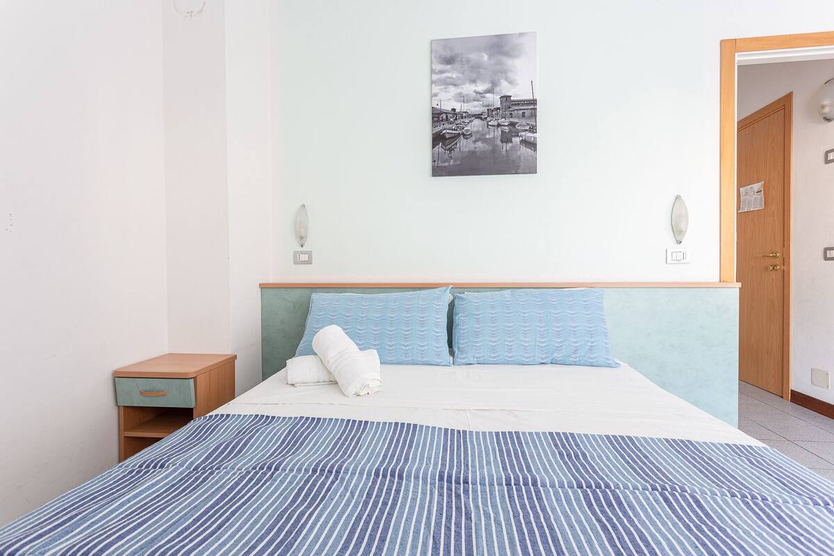 A neatly made bed with a striped bedspread, a headboard with a light blue panel, and a wall-mounted picture of a harbor scene.