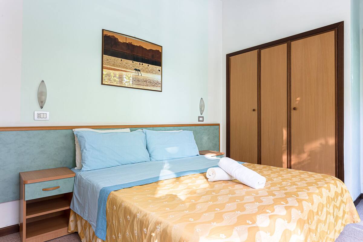 A neatly made bed with a patterned bedspread, a nightstand with a drawer, a wall-mounted picture, and a wardrobe.