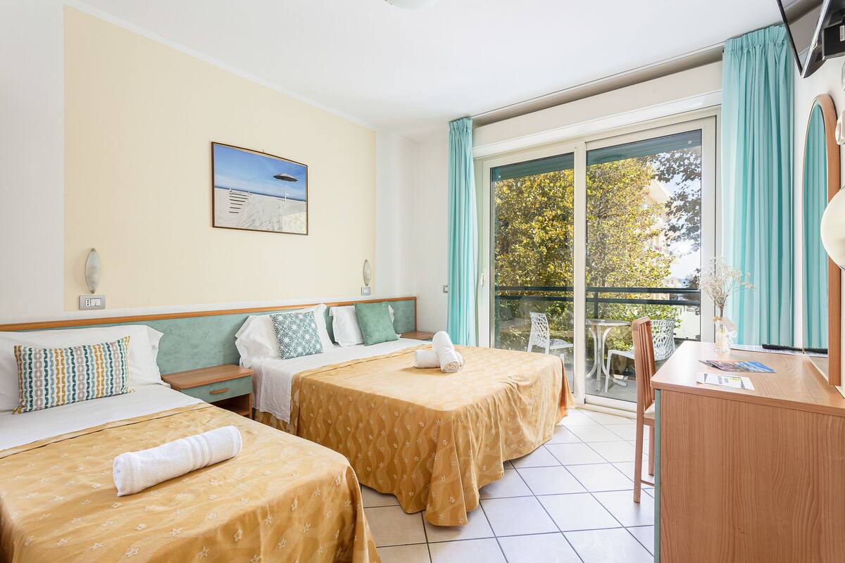 A hotel room with two beds, a desk, and a balcony with a view of trees.