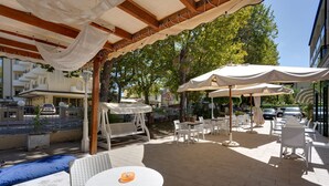 An outdoor patio area with white wicker furniture, including tables and chairs, under a wooden pergola with white fabric drapes. - Gadames Hotel - Lunch on the Beach (Cervia)
