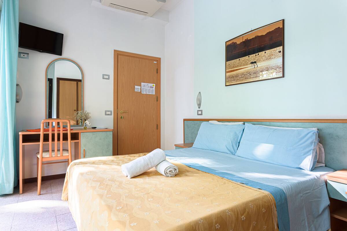 A hotel room with a bed, a desk, a chair, a television, and a painting on the wall.