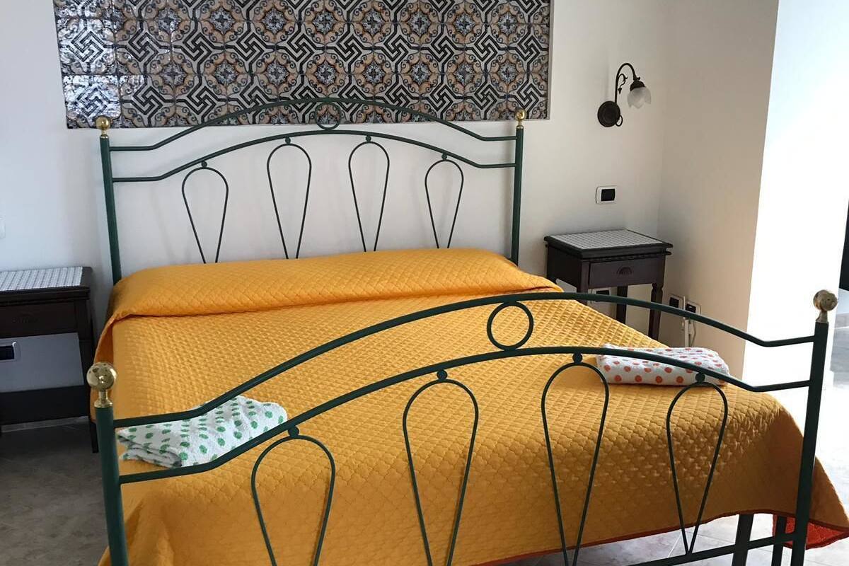 Ein Bett mit Metallgestell und einer gelben Steudecke, eine Wand mit Wandteppichmuster und ein kleiner Nachttisch mit Lampe.