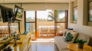 A modern living room with a sofa, a coffee table, and a TV. There is a balcony with a view of palm trees and a building. - Cobertura com Piscina Privativa | Pé na Areia | Vista Mar | Barra Bali | Destino BSM 417 (Barra de São Miguel)