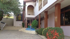 A building with a covered entrance, steps leading up to it, and a small garden area with bushes and flowers. - Queensland Hotel (Sekondi-Takoradi)