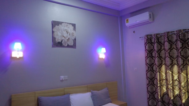A bedroom with a bed, two wall-mounted lamps, an air conditioner, and a patterned curtain. - Queensland Hotel (Sekondi-Takoradi)