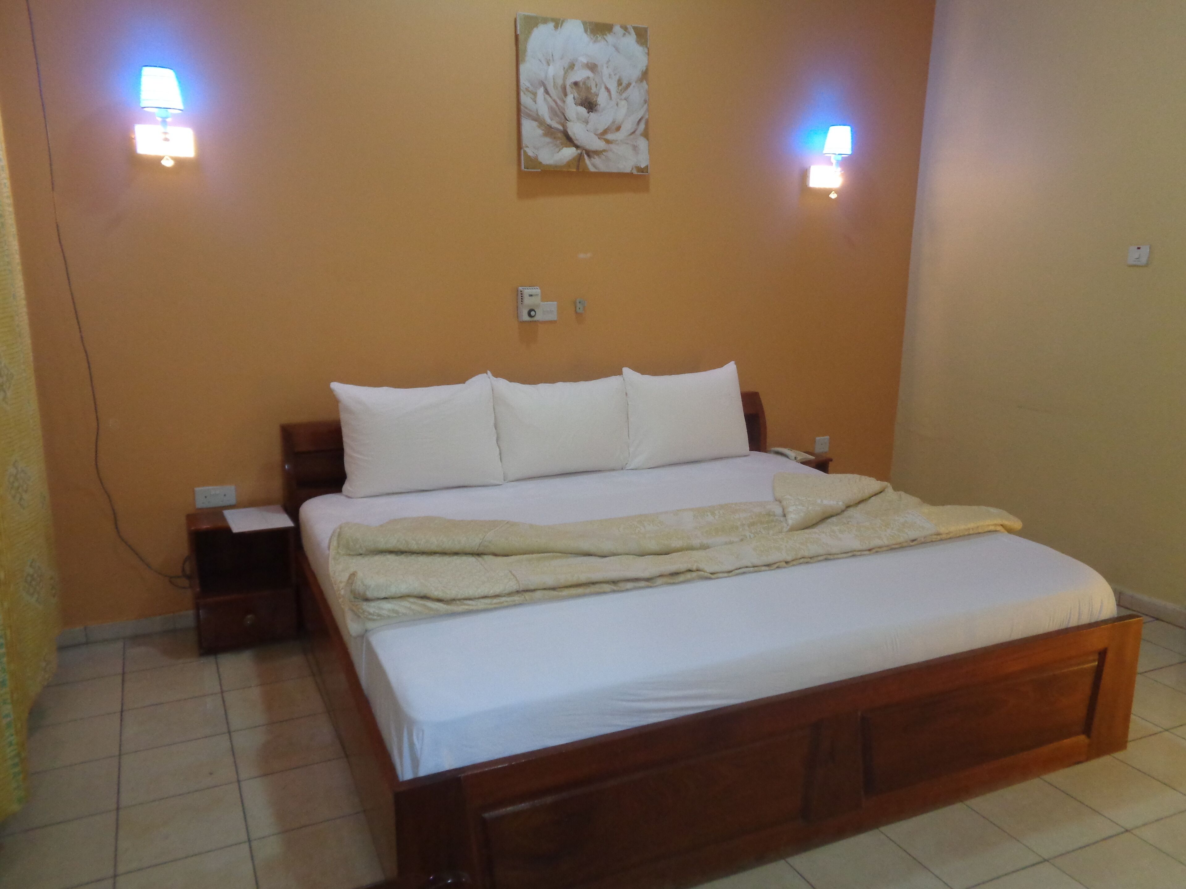 Una cama bien hecha con sábanas blancas y una manta color beige, dos mesitas de noche con lámparas, una lámpara de pared y un cuadro floral sobre la cama.