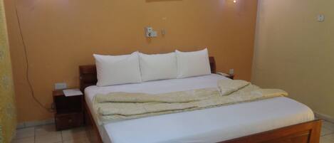 Una cama bien hecha con sábanas blancas y una manta color beige, dos mesitas de noche con lámparas, una lámpara de pared y un cuadro floral sobre la cama.