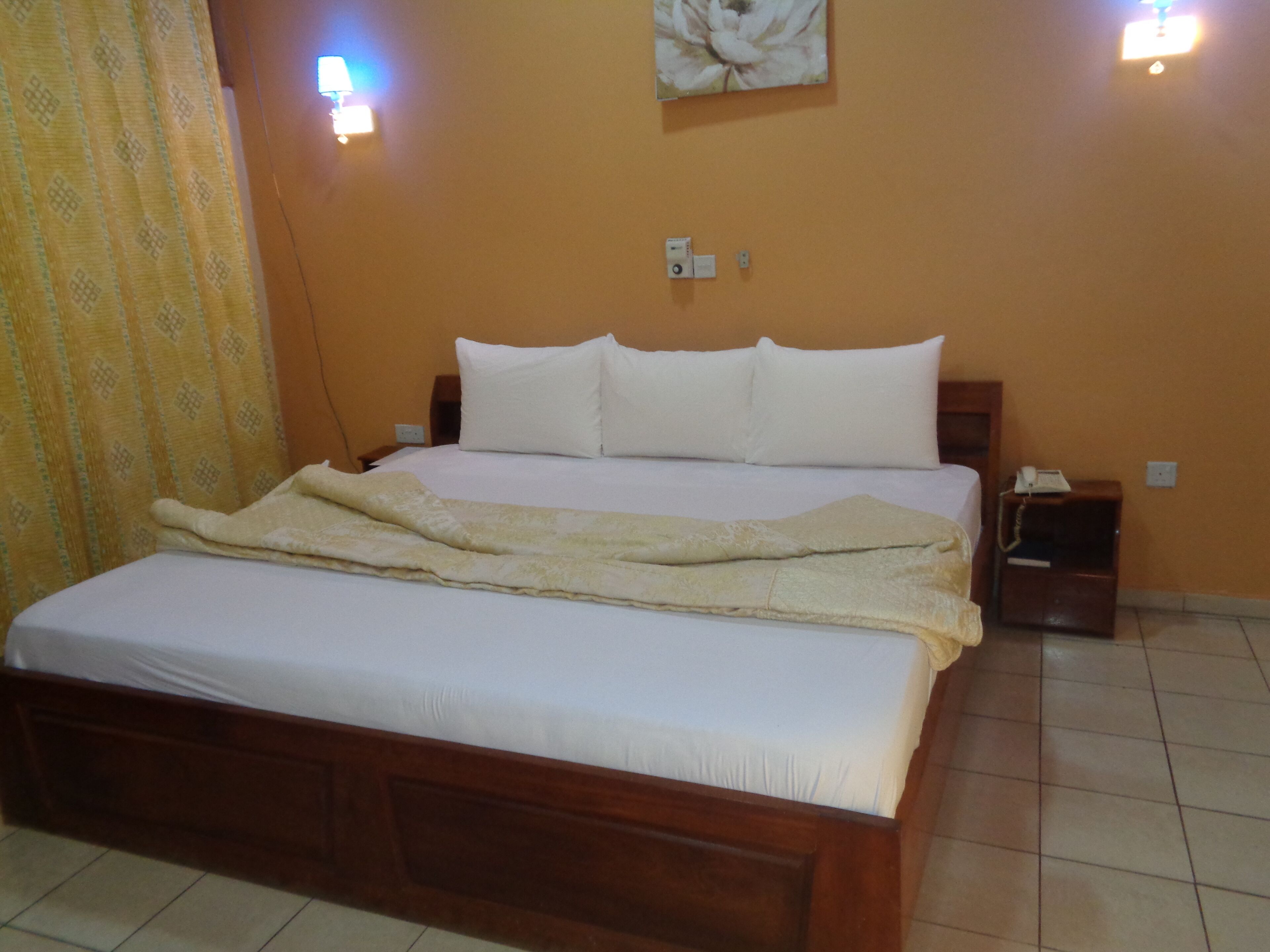 Cama bem arrumada com roupa de cama branca e manta bege, cabeceira de madeira, mesa de cabeceira com telefone e luminária fixada na parede.