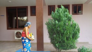 A statue of a woman holding a child and a basket, placed on a blue tiled platform outside a building with windows and a tree. - Queensland Hotel (Sekondi-Takoradi)
