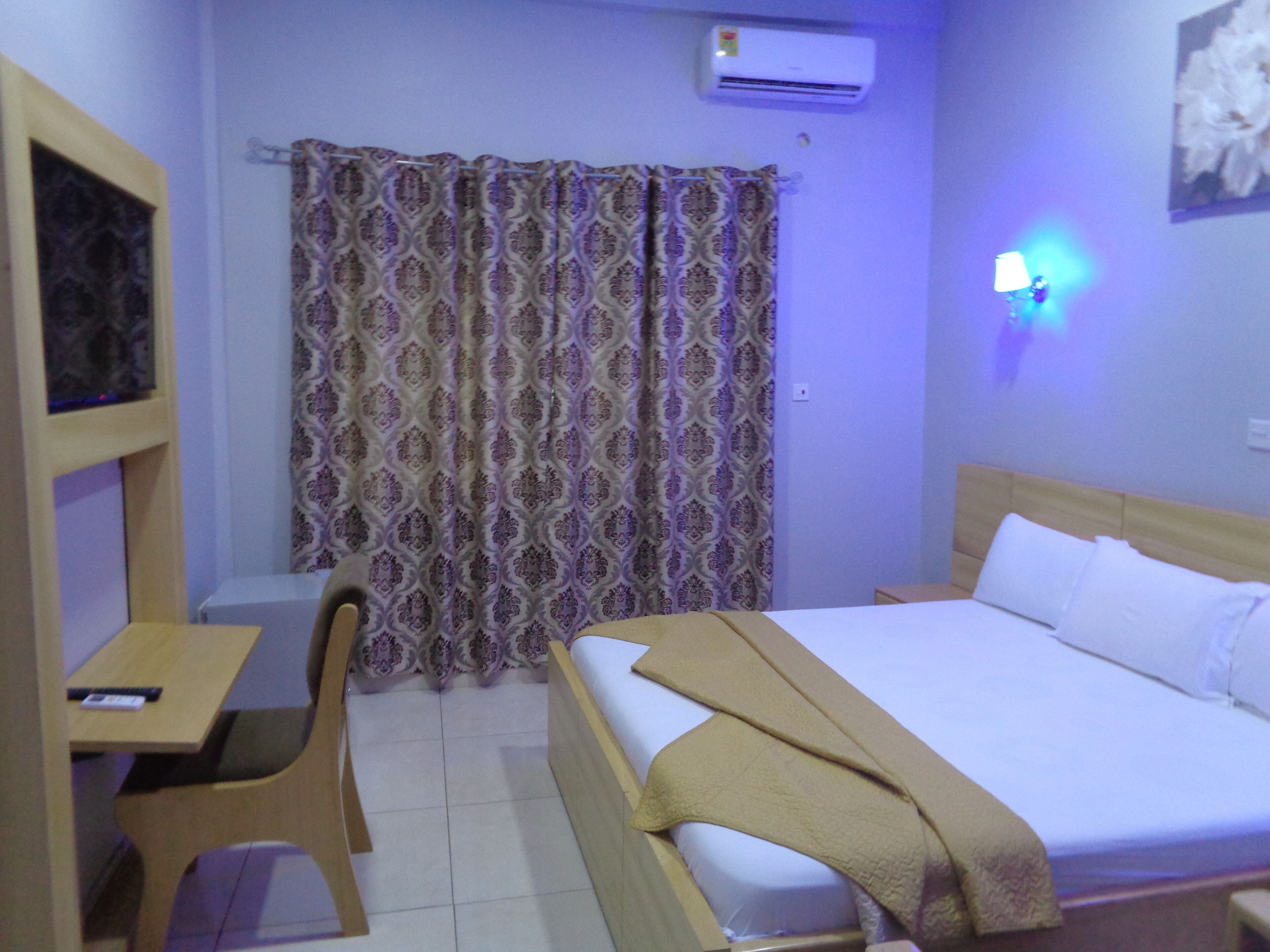 A hotel room with a bed, a desk, a chair, a curtain, and a wall-mounted lamp.