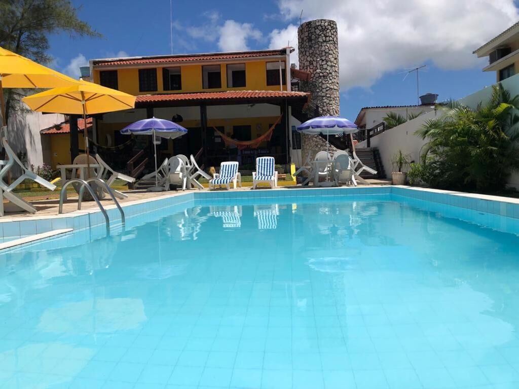 Uma piscina com espreguiçadeiras e guarda-sóis, um pilar de pedra e um edifício com telhado vermelho ao fundo.