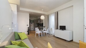 Un salon moderne avec un canapé, des coussins verts, un fauteuil jaune, un coin repas avec une table et des chaises, une cuisine en arrière-plan et un téléviseur posé sur un meuble blanc.