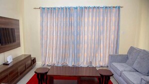 Sala de estar con un televisor de pantalla plana montado en un panel de madera, un sofá gris, una mesa de centro de madera y una cortina estampada.