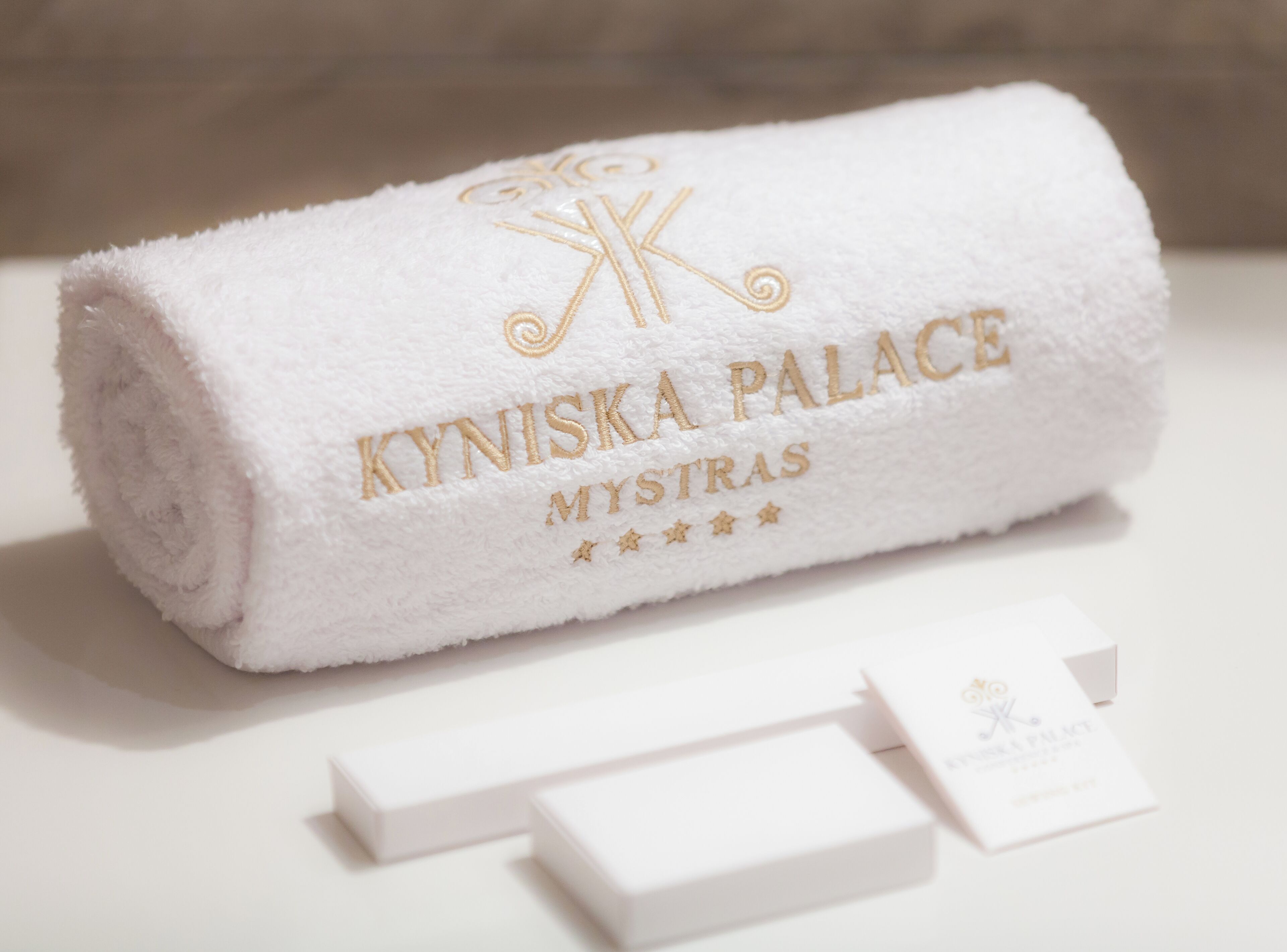 A white towel with embroidered text "KYNISKA PALACE MYSTRAS" and a logo, placed on a white surface with two white cards beside it.