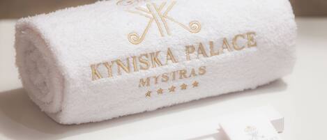 A white towel with embroidered text "KYNISKA PALACE MYSTRAS" and a logo, placed on a white surface with two white cards beside it.