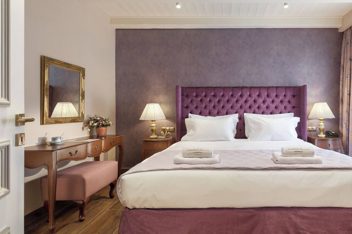 A hotel room with a large bed, two bedside tables with lamps, a wooden desk, and a bench.
