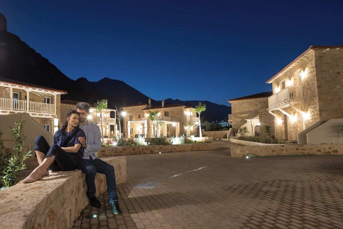 A couple sitting on a stone bench in a courtyard with stone buildings and a mountain in the background.