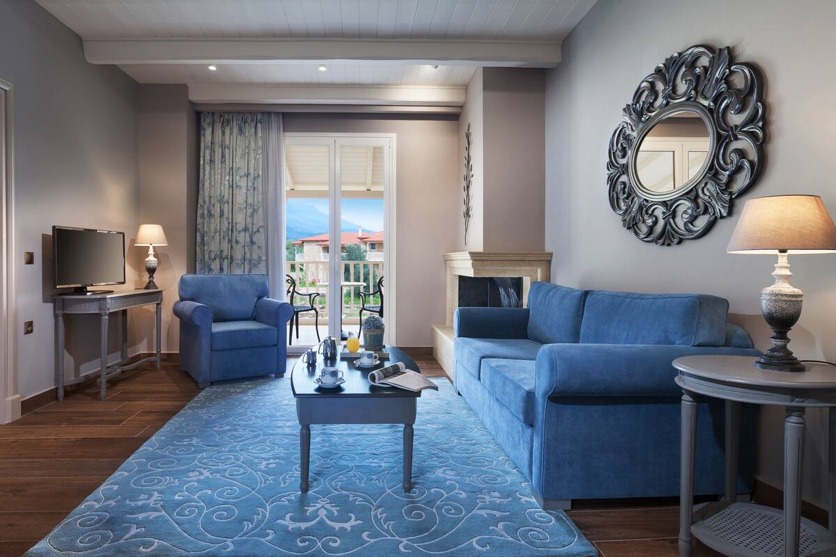 A hotel room with a bed, a desk with a TV, a blue armchair, a sofa, a coffee table, and a view of the outside.