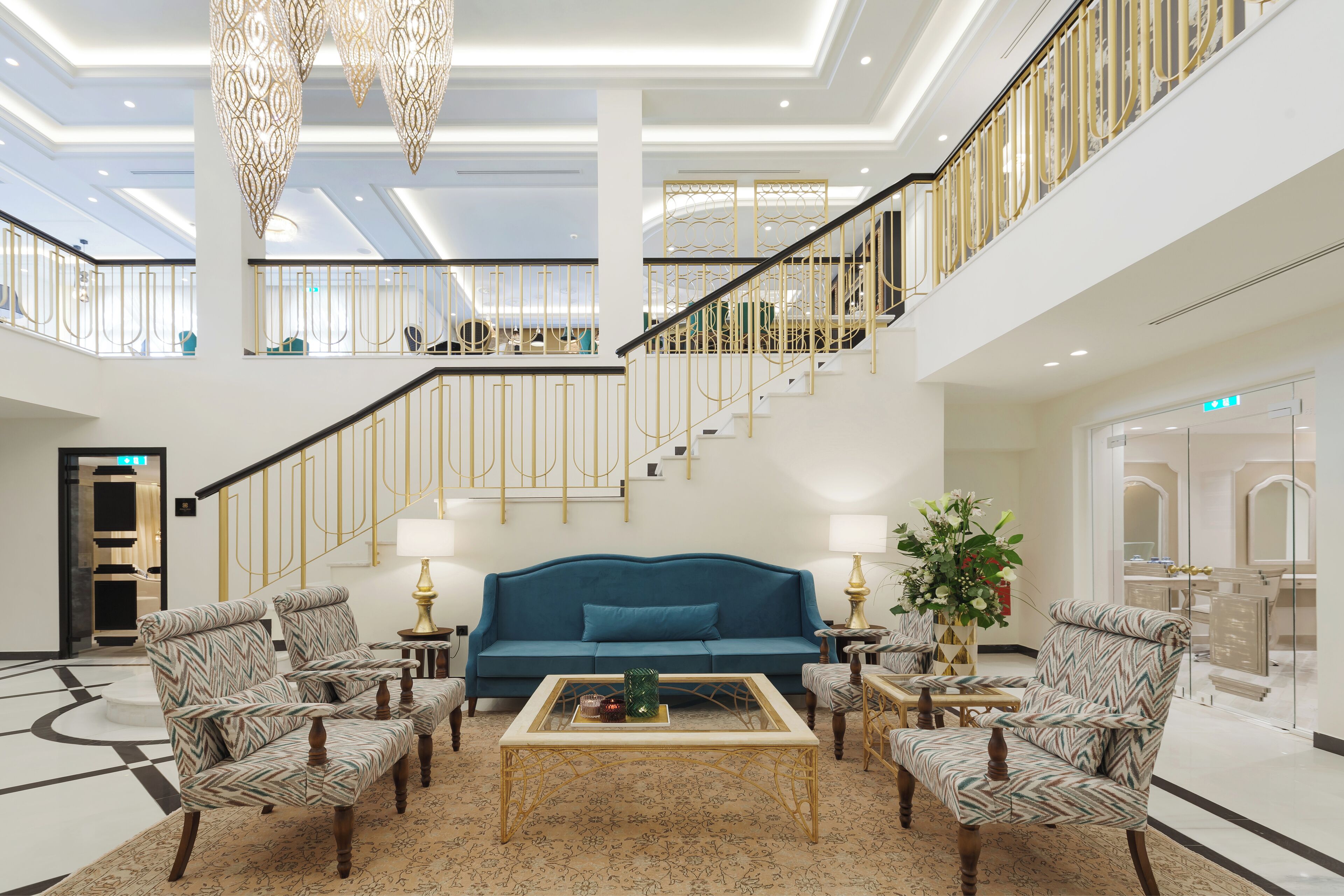 A spacious lobby with a blue sofa, patterned armchairs, a coffee table, and a staircase with golden railings.