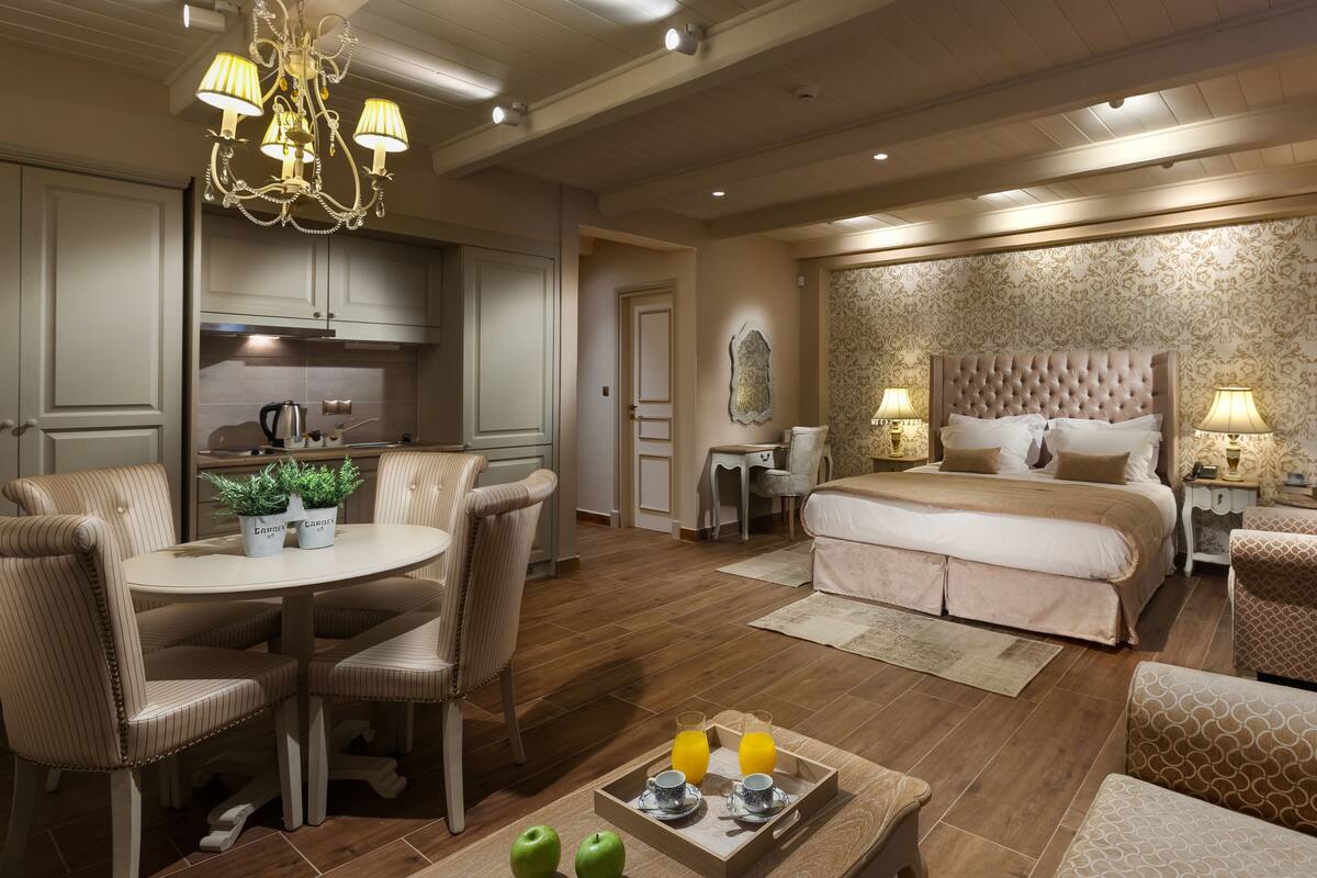 A hotel room with a dining area, a bed with a large headboard, a desk, and a chandelier.