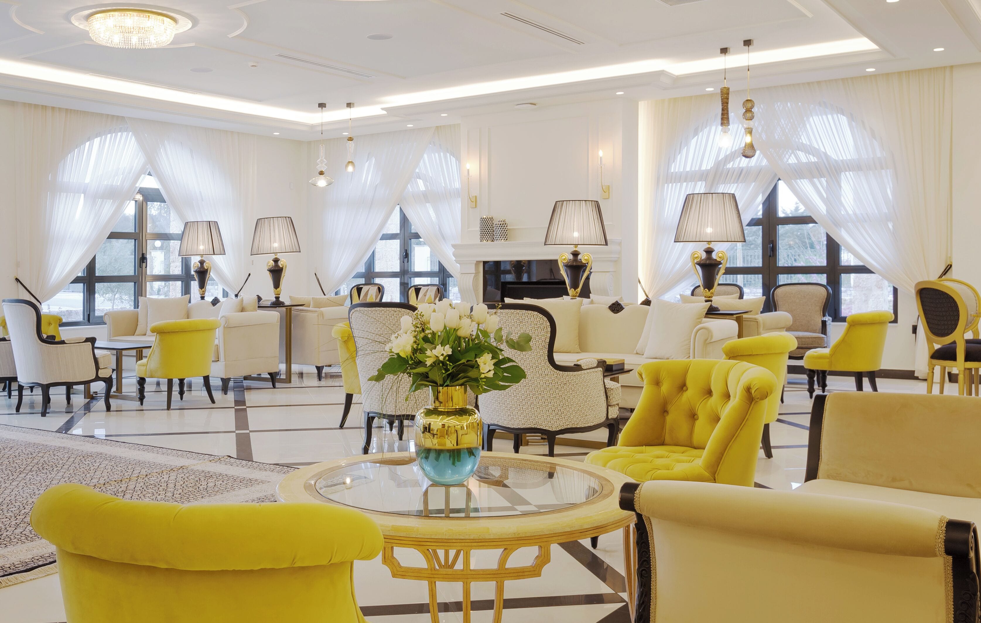 A spacious, elegantly furnished living room with a large round table, yellow armchairs, and a fireplace.