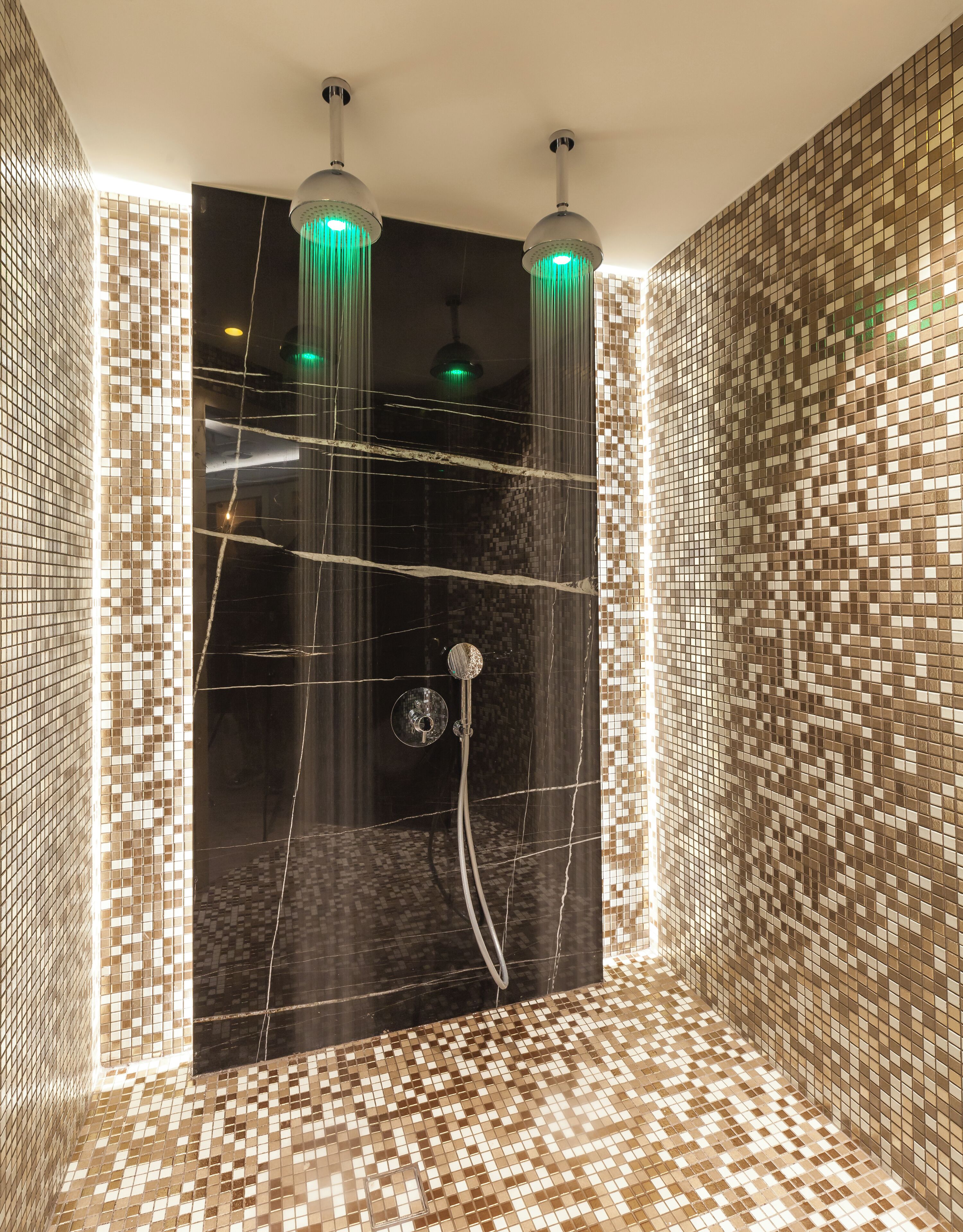 Un baño moderno con paredes y suelo de mosaicos, zona de ducha con mampara de vidrio y dos luminarias colgantes.