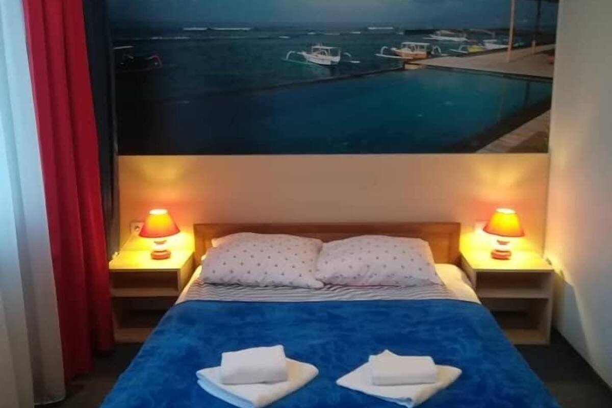 A hotel room with a bed, two bedside lamps, a red curtain, and a large painting of a marina.