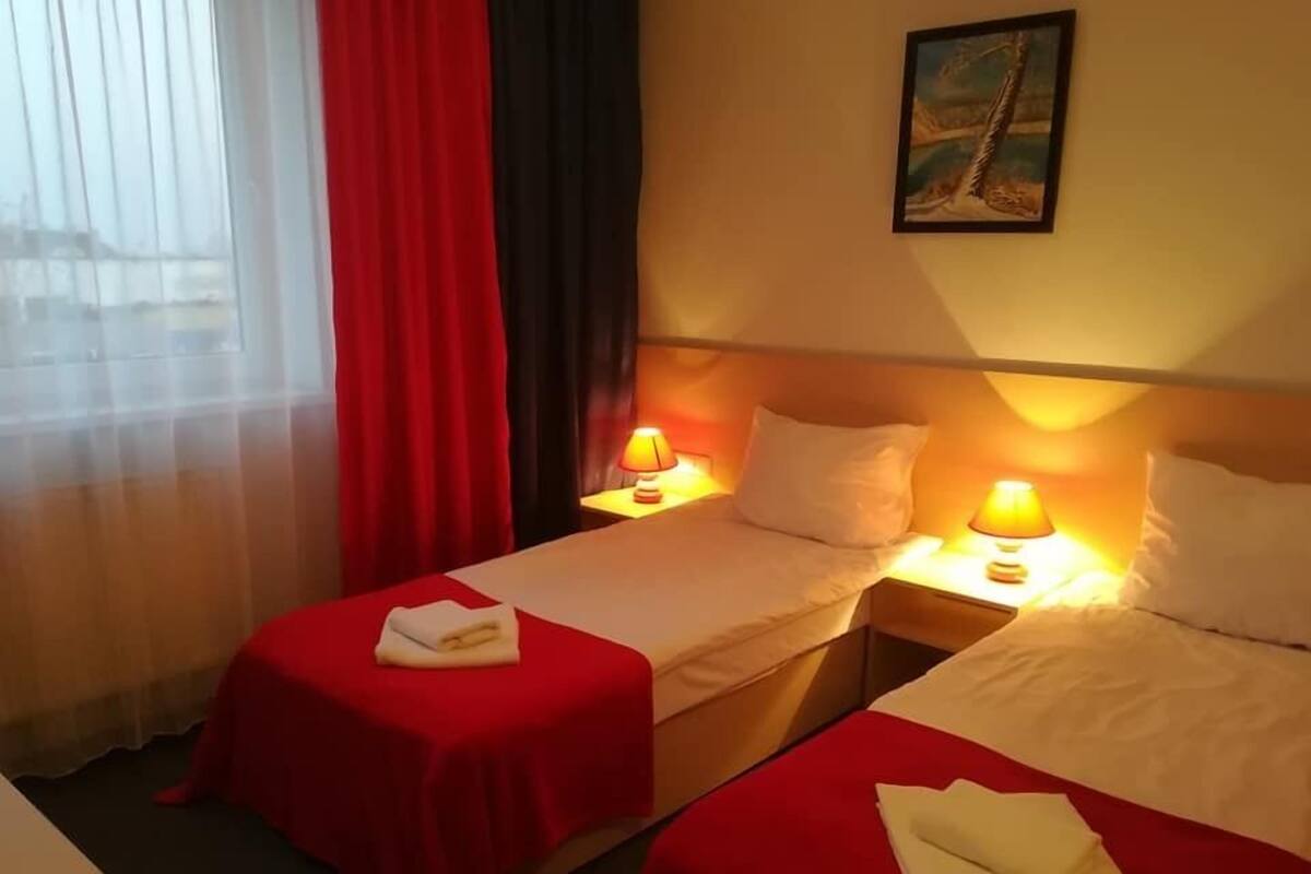 A hotel room with two beds, each with a red bedspread and white pillows. There are two bedside lamps, and a painting is visible on the wall.