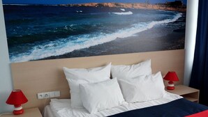 Una habitación de hotel con cama, almohadillas blancas, colcha roja y azul marino, lámpara roja en la mesita de noche y un mural en la pared con vista al océano.