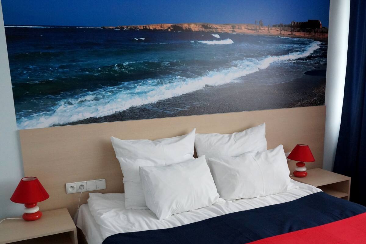 A hotel room with a bed, white pillows, a red and navy bedspread, a red lamp on the nightstand, and a wall mural of the ocean.