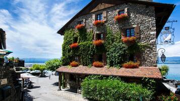 Un bâtiment en pierre, agrémenté de jardinières rouges aux fenêtres, doté d’une terrasse et offrant une vue sur l’eau.