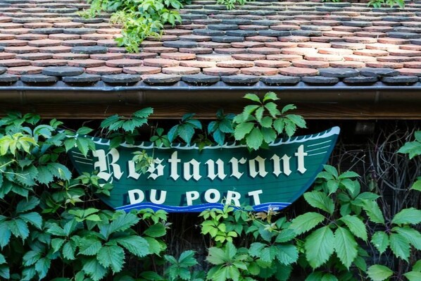 A sign for a restaurant named "Restaurant du Port" with a green background and white lettering, partially obscured by green foliage.