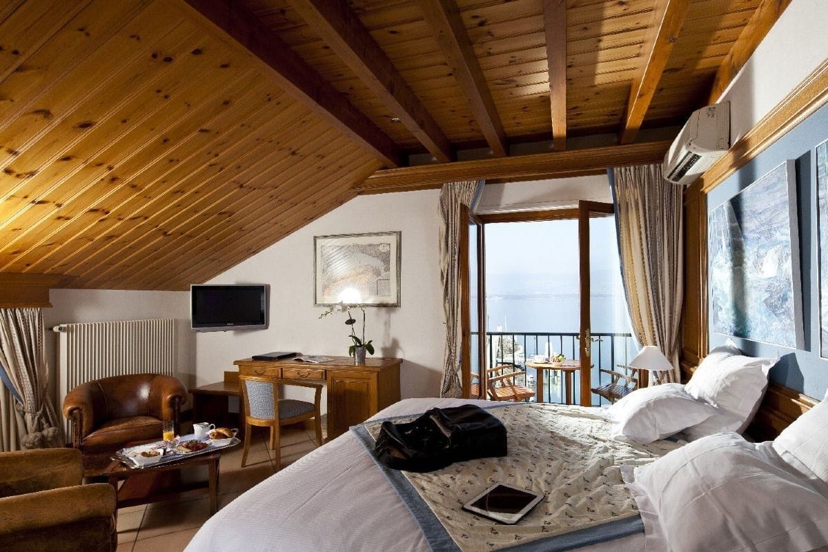 Una habitación de hotel acogedora con techo de madera, cama con ropa blanca, escritorio con silla, televisión y balcón con vistas.