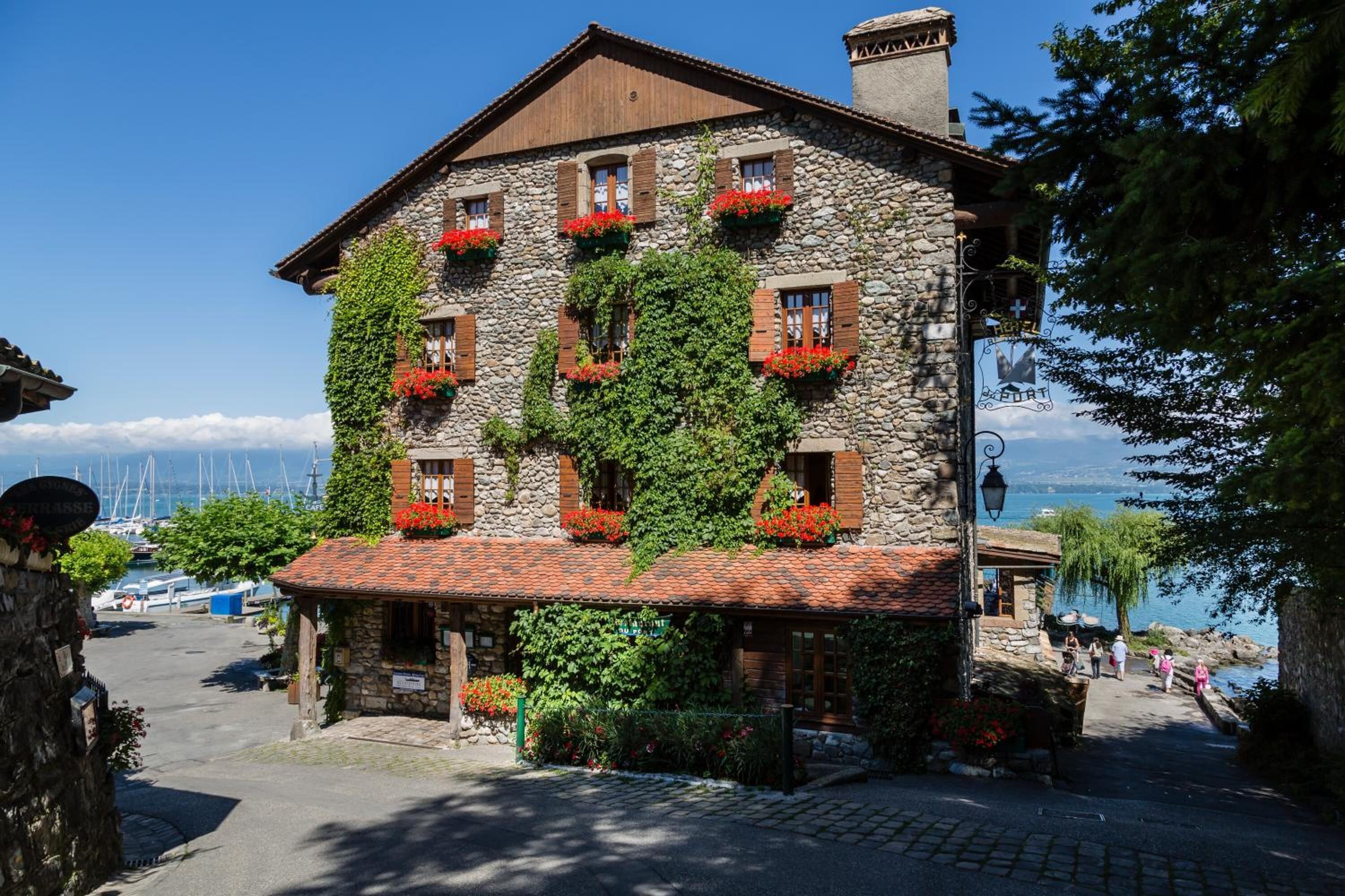 A stone building with red shutters and green vines, located near a body of water with boats.