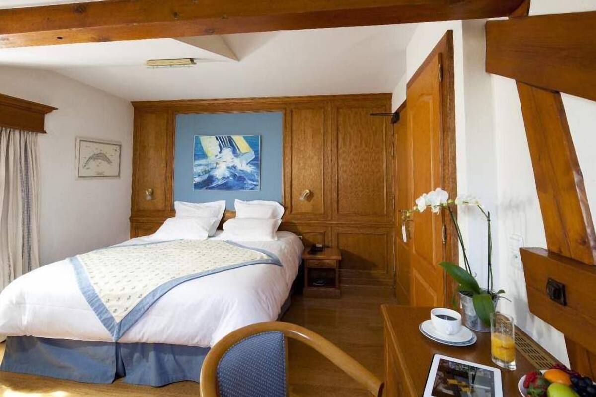 Habitación de hotel con una cama grande, cabecera de madera, un cuadro en la pared, una mesita con una taza y fruta, y una silla.