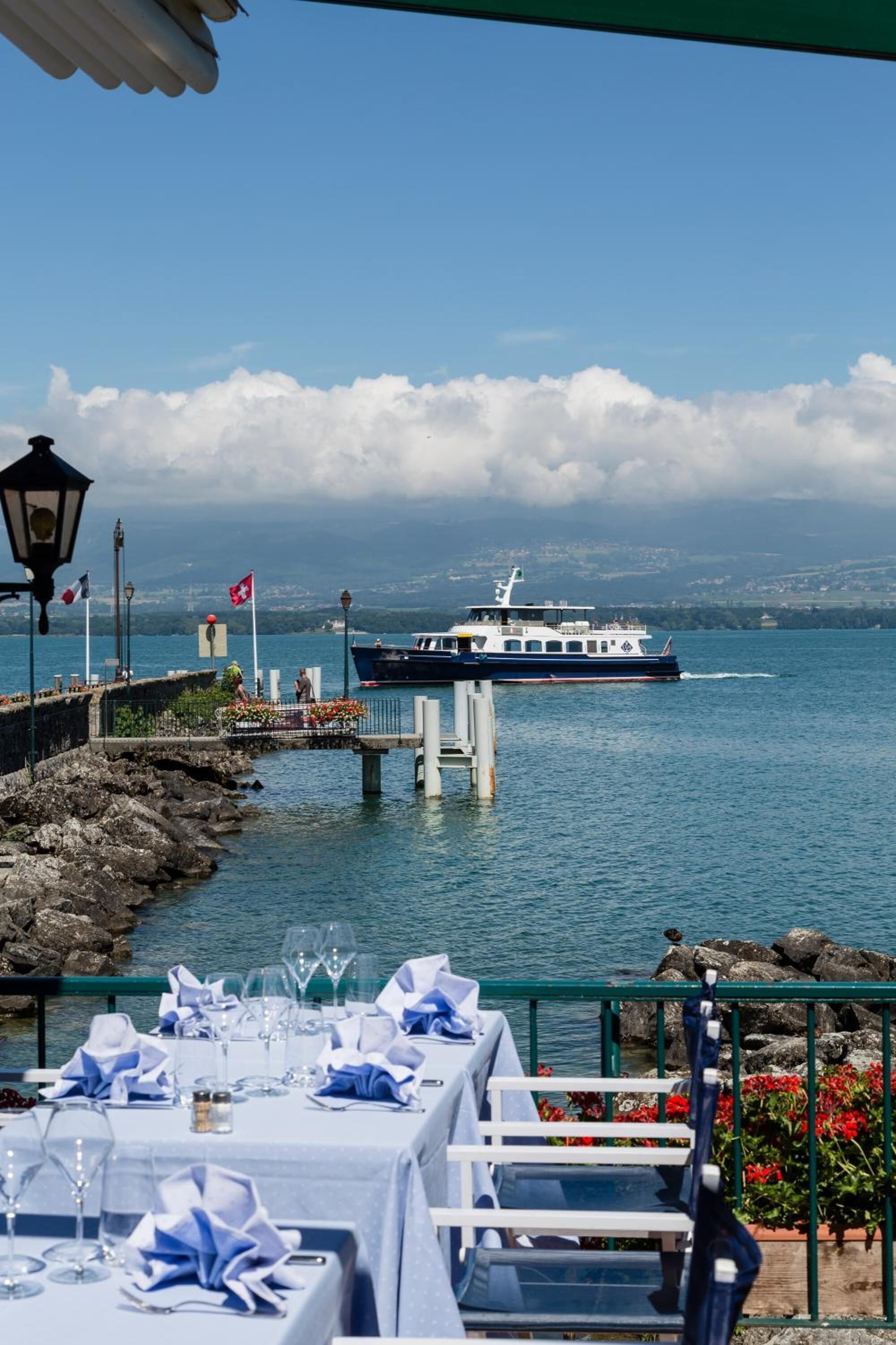A waterfront restaurant with tables set for dining, overlooking a lake with a boat in the distance.