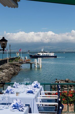 Een restaurant aan het water met gedekte tafels, met uitzicht op een meer en een boot in de verte.