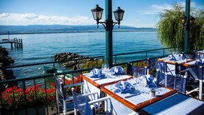 Un área de comedor al aire libre con mesas preparadas para dos, con vista a una masa de agua y una costa rocosa, y una cadena montañosa distante.