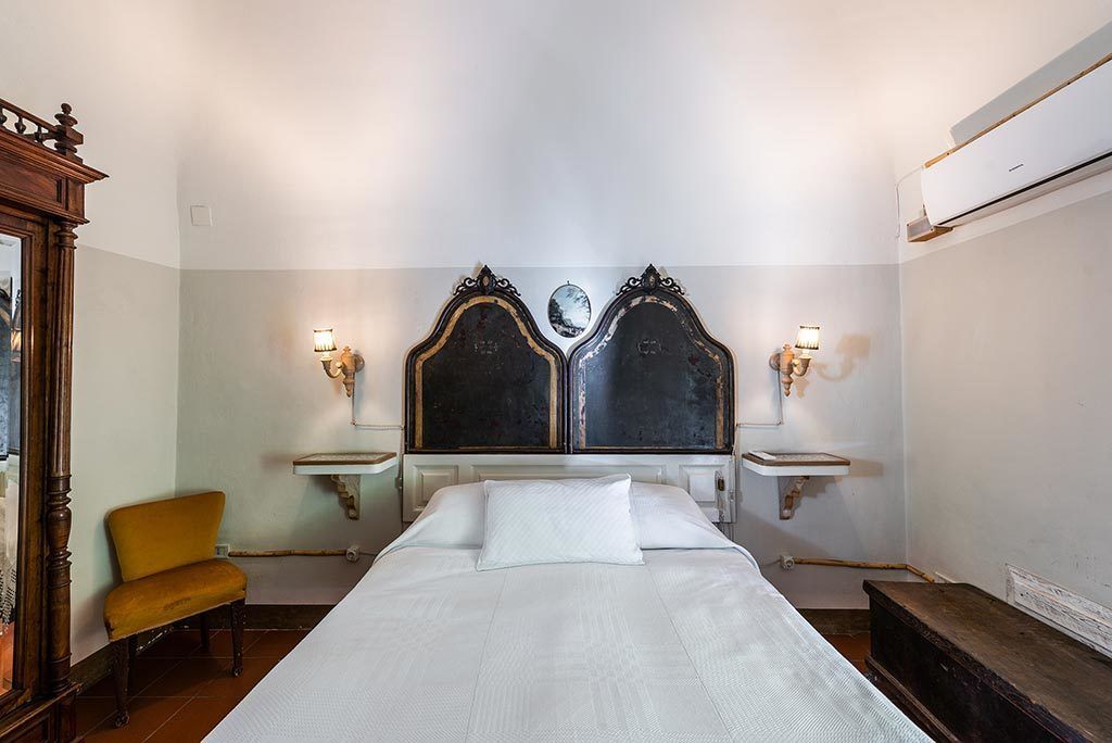 Quarto de hotel com uma cama grande, duas mesas de cabeceira com abajures, uma cadeira de madeira e um espelho à esquerda.