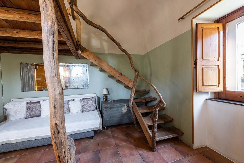 A rustic bedroom with a wooden staircase, a bed with white linens, a nightstand, and a window with curtains.