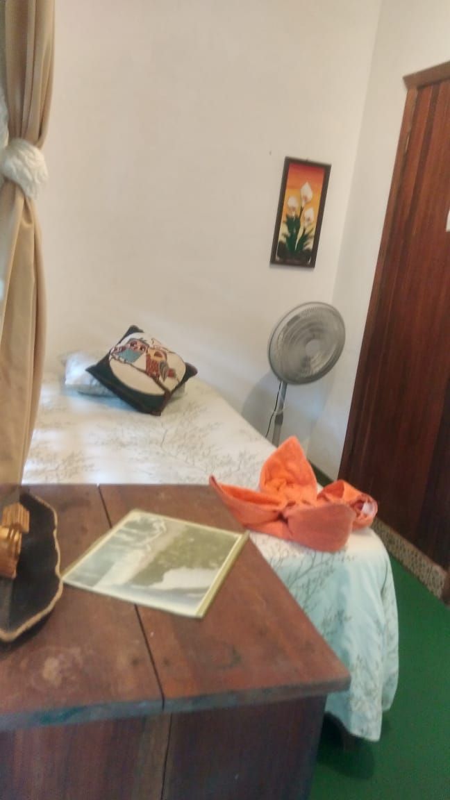 Ein Zimmer mit einem Bett, einem Nachttisch, einem Ventilator und einem Bild an der Wand.