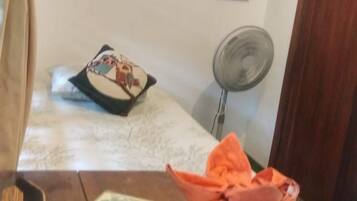 Ein Zimmer mit einem Bett, einem Nachttisch, einem Ventilator und einem Bild an der Wand.