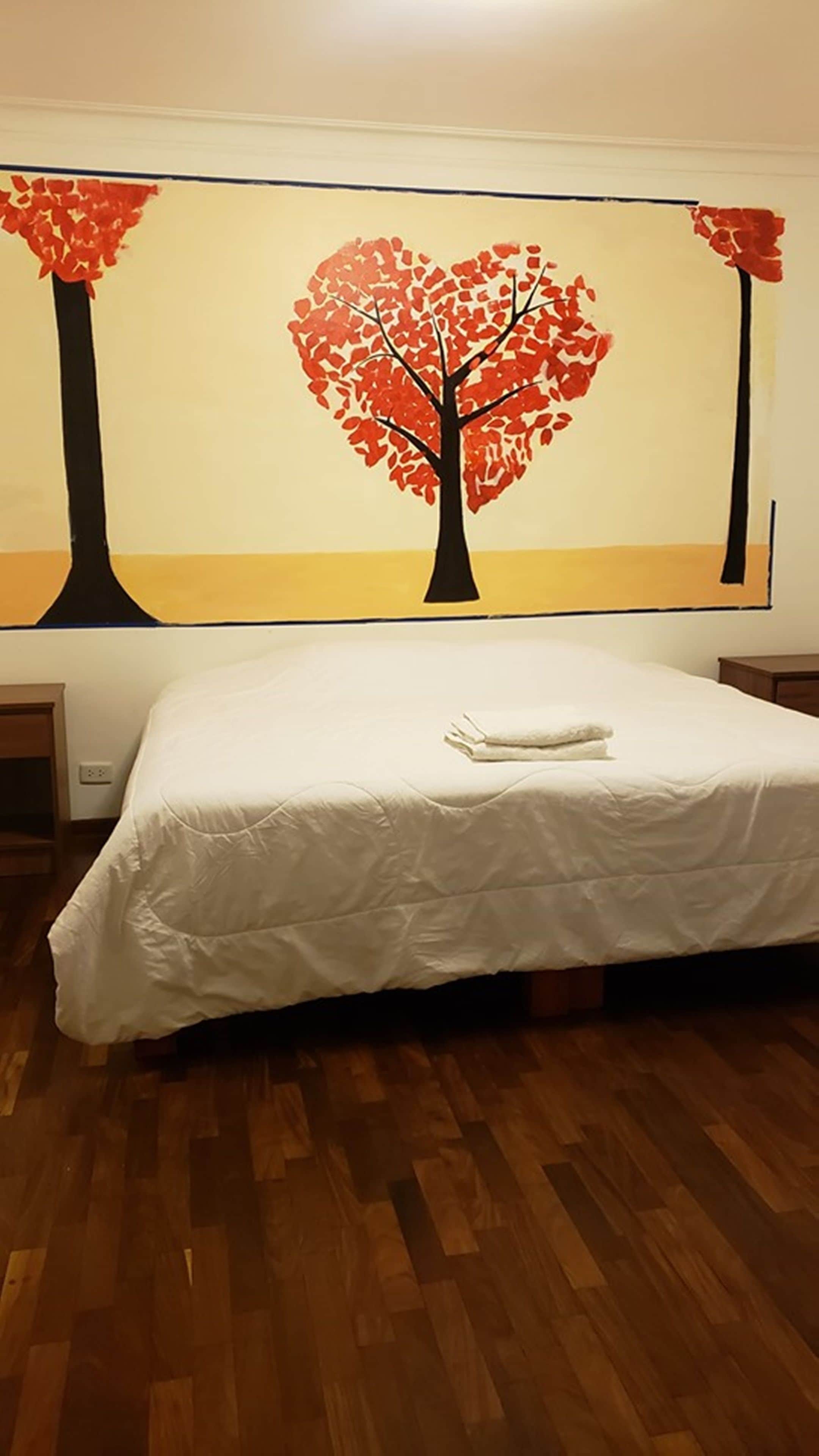 Hotellihuone, jossa on sänky, valkoinen sängynpeite ja seinämaalaus, jossa on kuvattuna puu punaisine lehtineen.